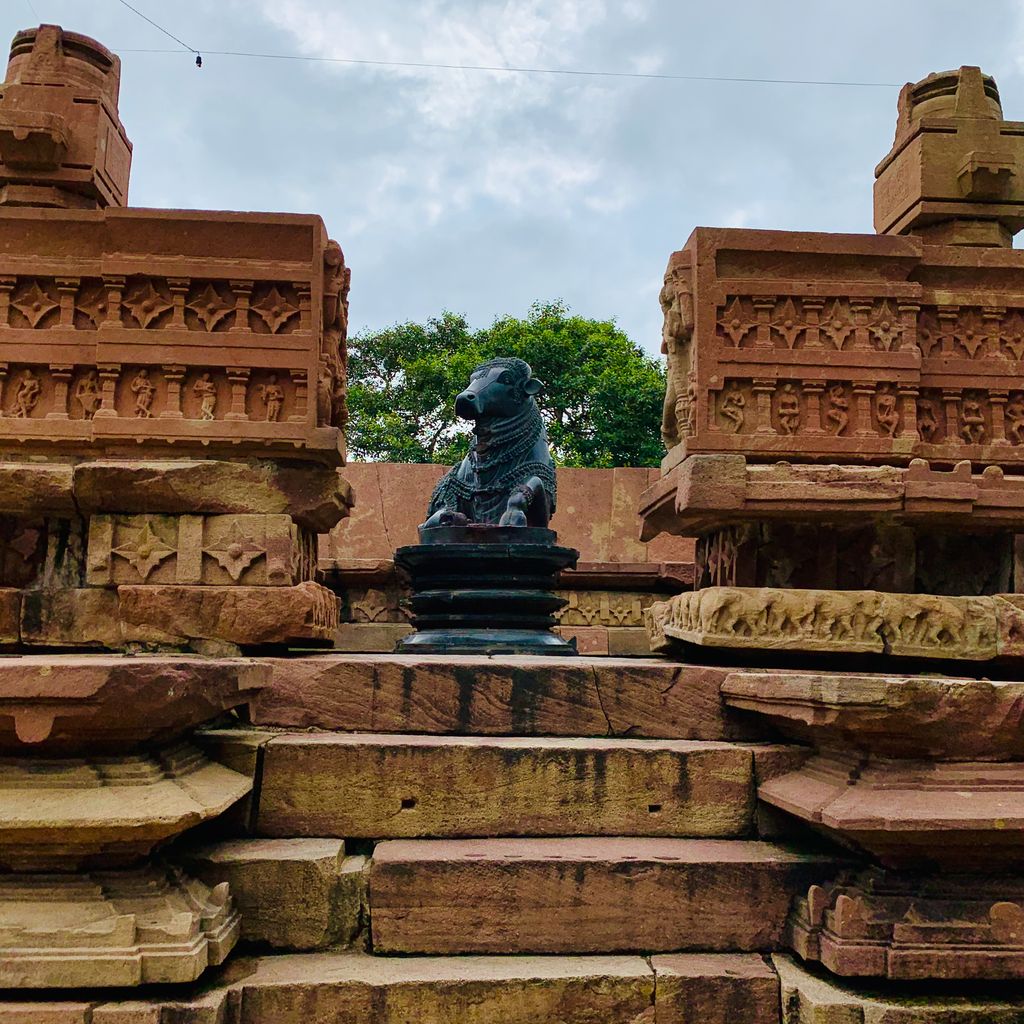 Stone steps rise between carved enclosure blocks towards a seated Nandi, with broken upper supports open to the sky.