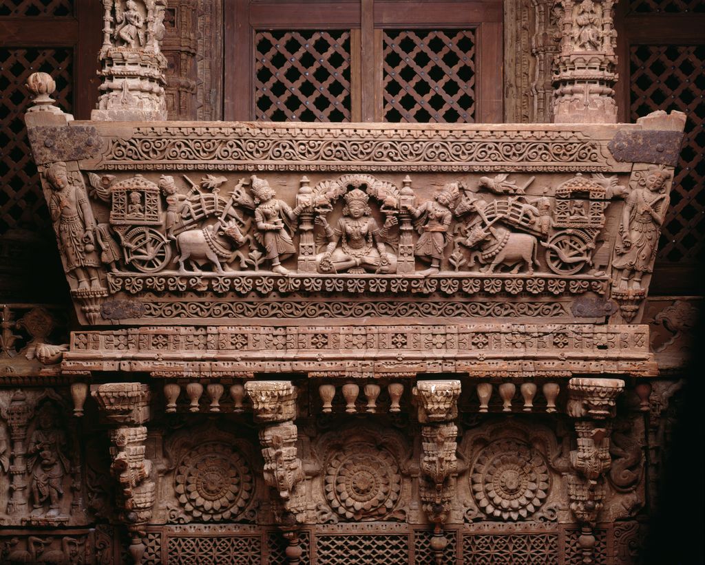 Densely carved wooden balcony members show figures, elephants and floral bands above small brackets, with lattice grating behind.