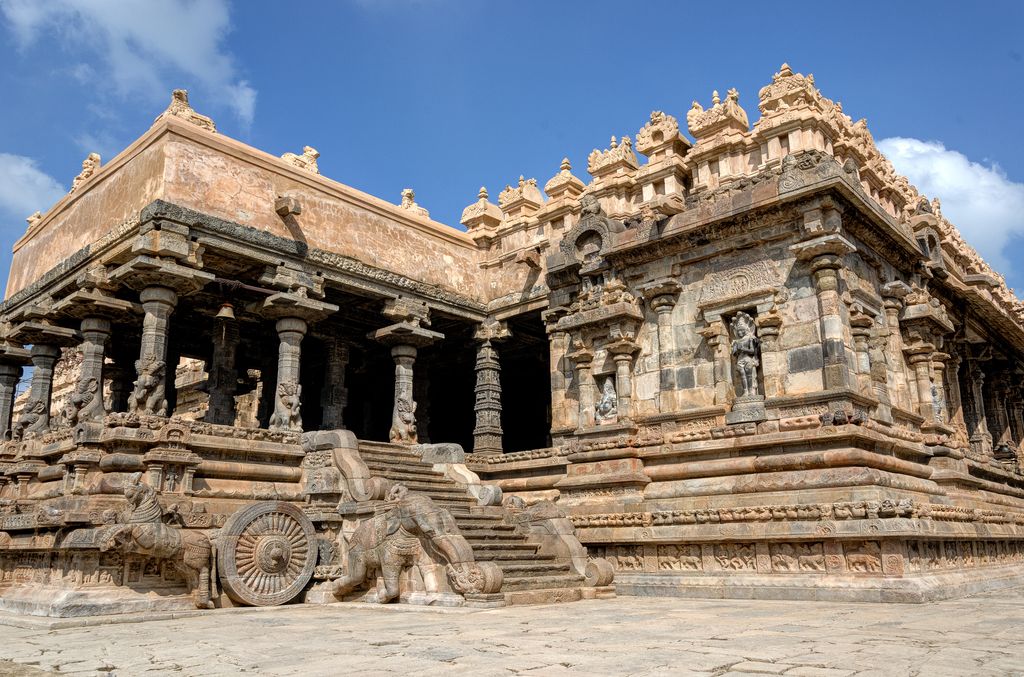 A carved stone wheel and horse stand beside steps leading to an open columned porch, adjoining a richly carved temple wall.
