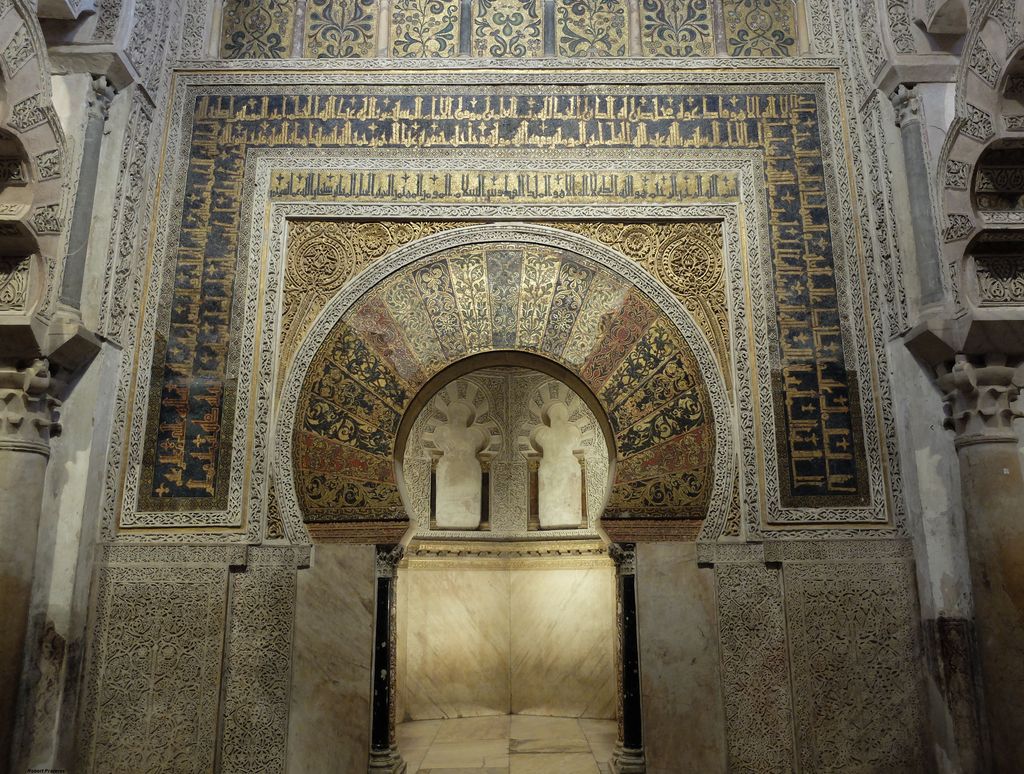 A gold and coloured mosaic horseshoe arch opens into a small marble-lined chamber with blind arches on its rear wall.
