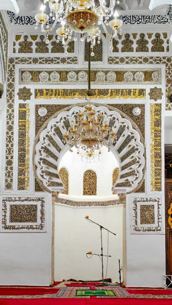 A white-and-gold ornamented frame surrounds a deep niche, with chandeliers above and microphone stands near prayer rugs below.