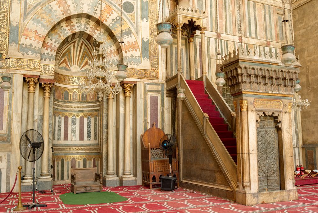 A decorated recessed niche stands beside a tall minbar with red-carpeted steps, within a marble-clad wall.