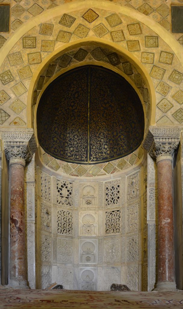 Pierced pale panels line a curved niche beneath a dark patterned hood, framed by red columns and diamond-set lustre tiles.