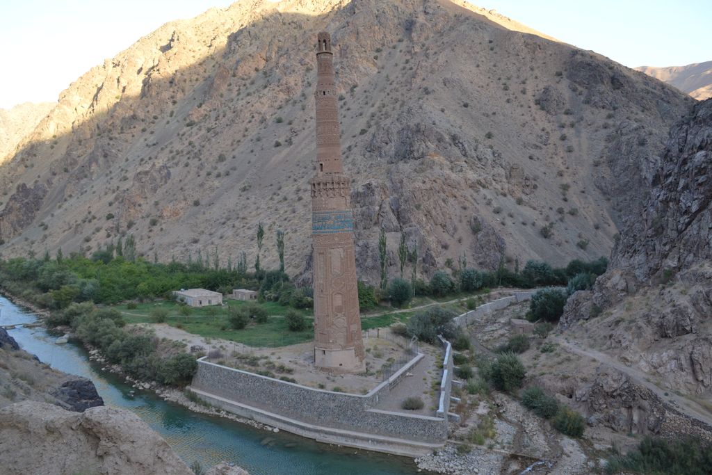 A slender patterned brick tower with a turquoise band stands beside a river beneath steep rocky slopes.