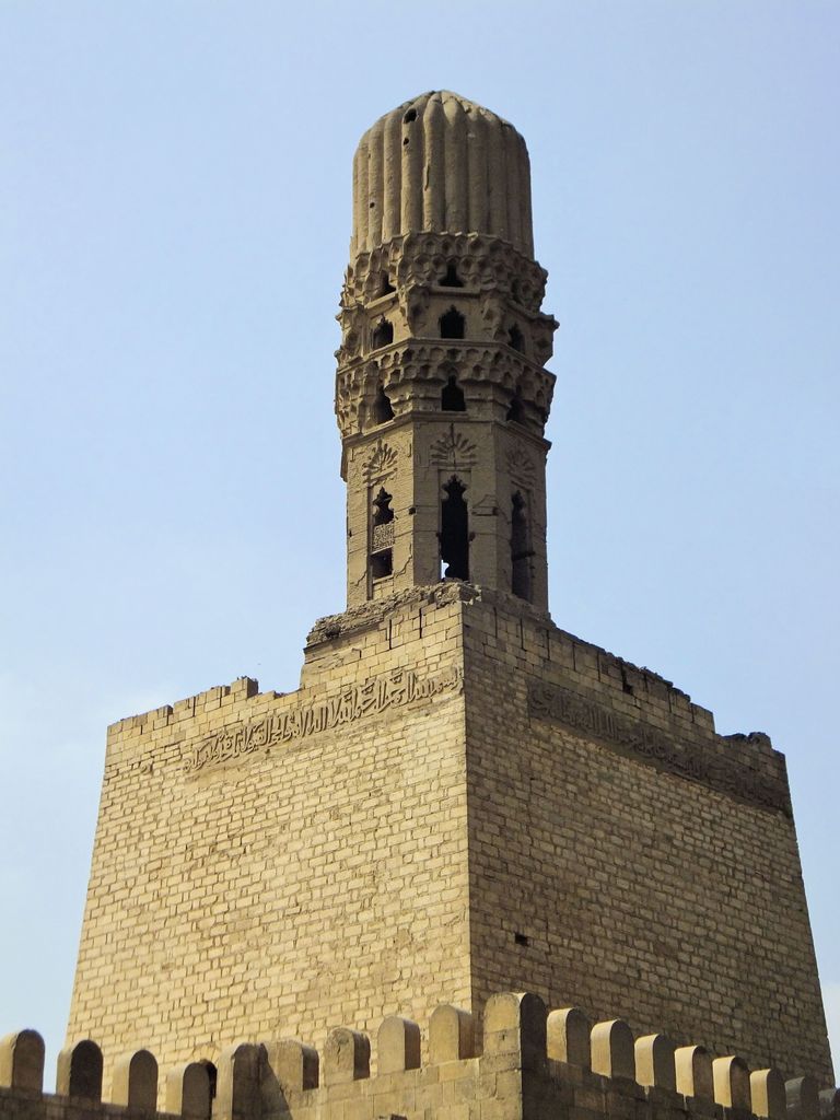 A large square masonry enclosure supports a narrow ornamented minaret crown above the crenellated city wall.