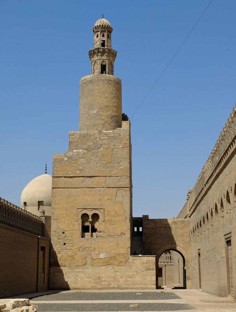 A square minaret base with external steps rises to a round upper stage and open crown, with an arched connection at right.
