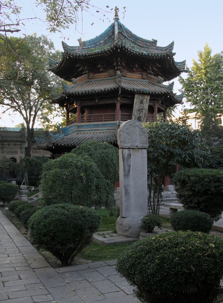 A timber pavilion with layered upturned tiled roofs rises behind a stone stele, clipped shrubs and trees.