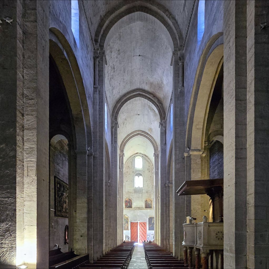 A slightly pointed barrel vault extends above narrow arcades and pews, with a central passage leading to red entrance doors.