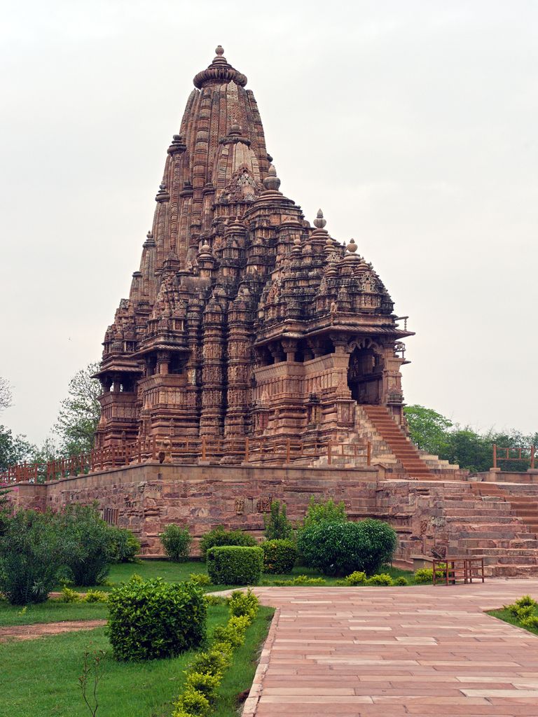 A tall curved stone spire rises through a cluster of smaller spires above stepped hall roofs, a high plinth and entrance steps.