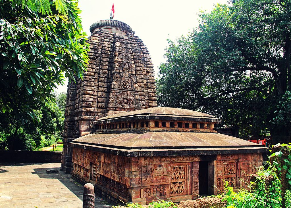 A curved carved tower with a ribbed crown and red flag rises behind a low stone hall with shallow sloping roof levels.