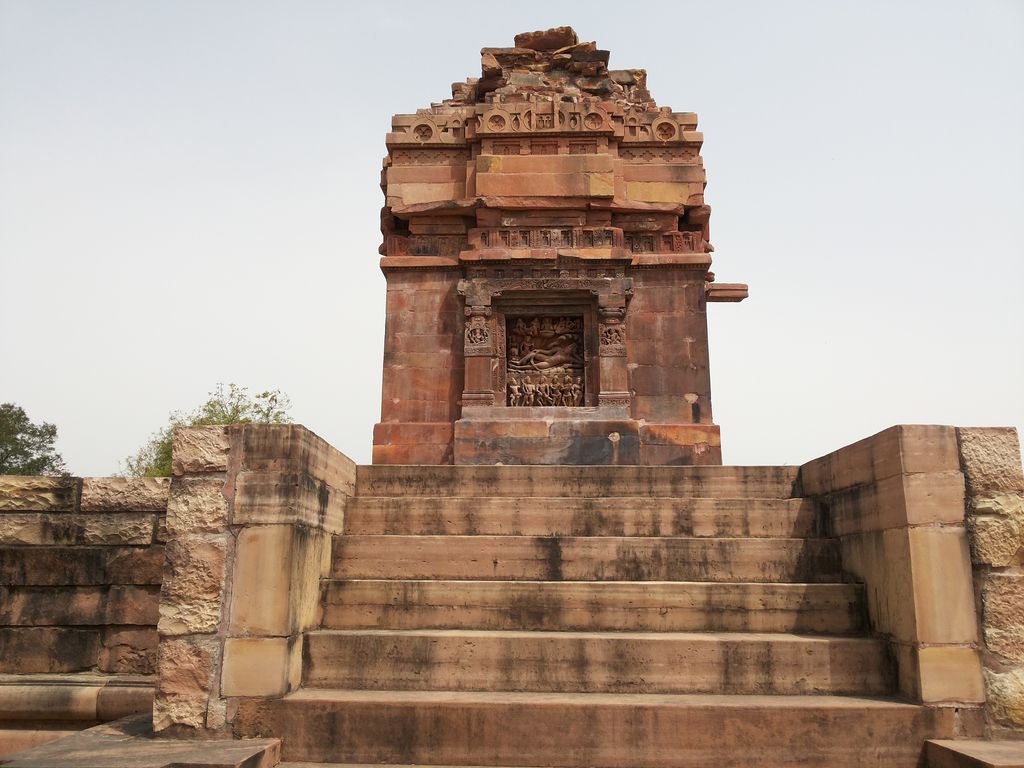 Steps rise towards a stone sanctuary wall with a carved relief, beneath incomplete stepped upper masonry.