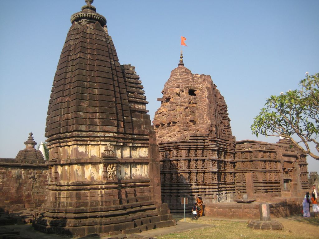 Carved stone shrines stand among trees and visitors; the central temple has uneven upper masonry, while a foreground shrine is cropped at the top.