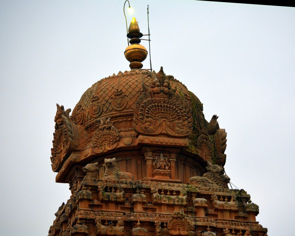 A faceted rounded stone cap supports a pot-shaped finial above carved arches and Nandi figures; a modern light is visible.