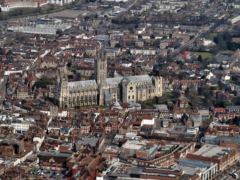 An elongated cathedral has a central tower and projecting transverse masses at different points along its roofs, surrounded by precinct buildings and city streets.