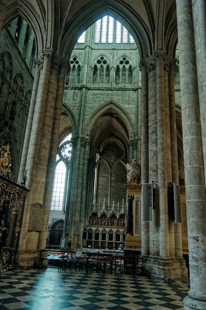 A tiled route passes between clustered piers and a screen into a tall vaulted transverse space with windows and chairs.