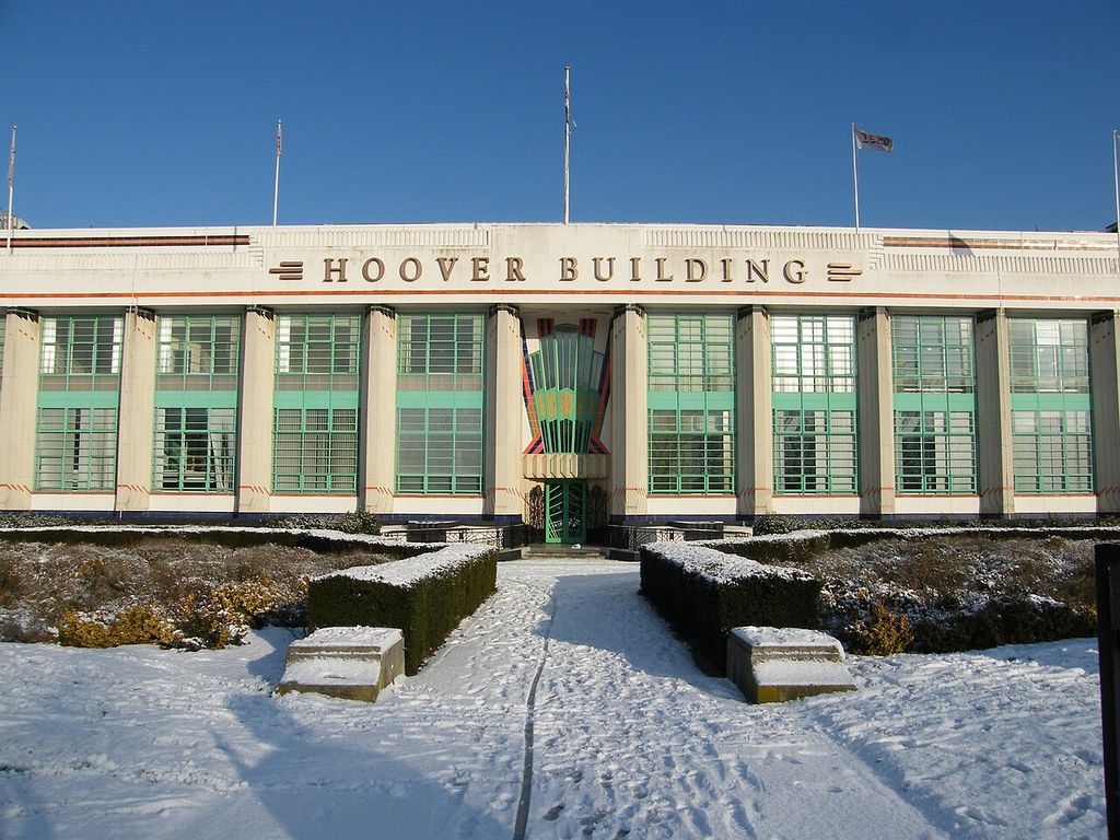 A long pale factory frontage has repeated green windows and a colourful fan-shaped central entrance, with snow in the garden.