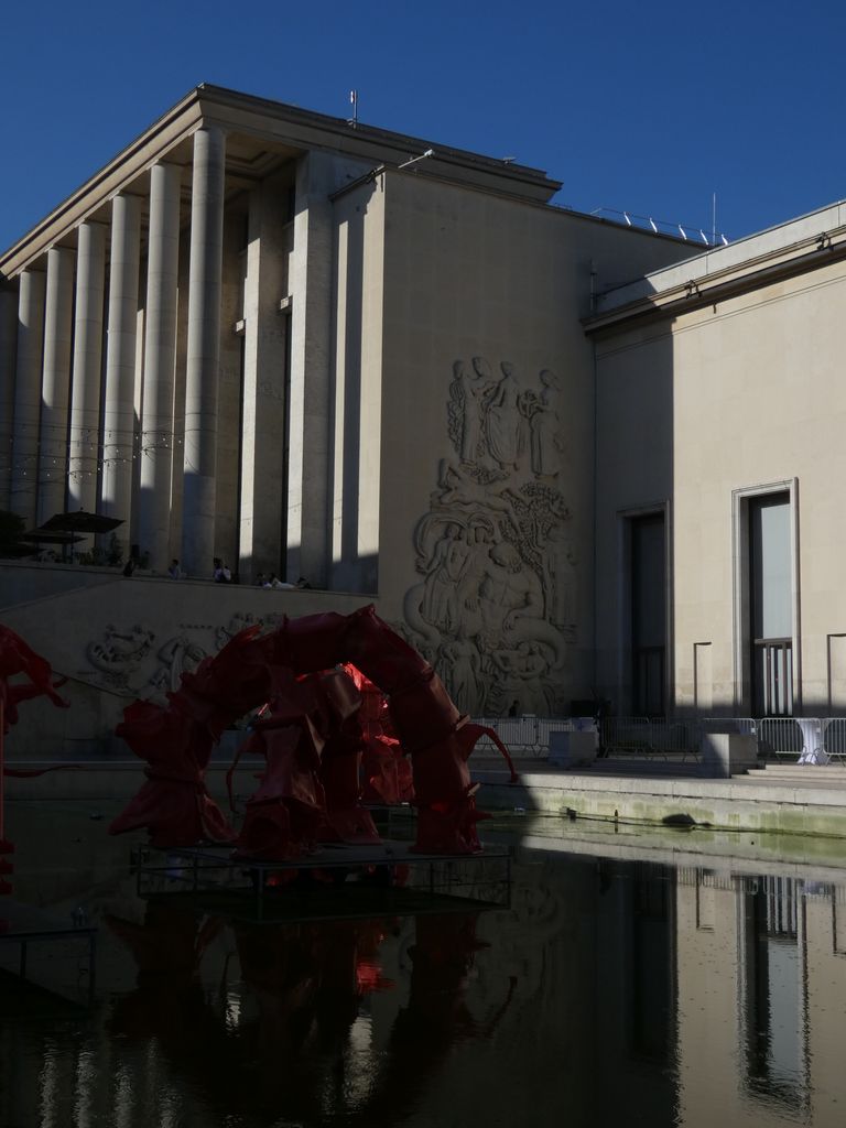 Tall plain columns rise beside a smooth wall with carved relief above a shadowed fountain and red artwork.