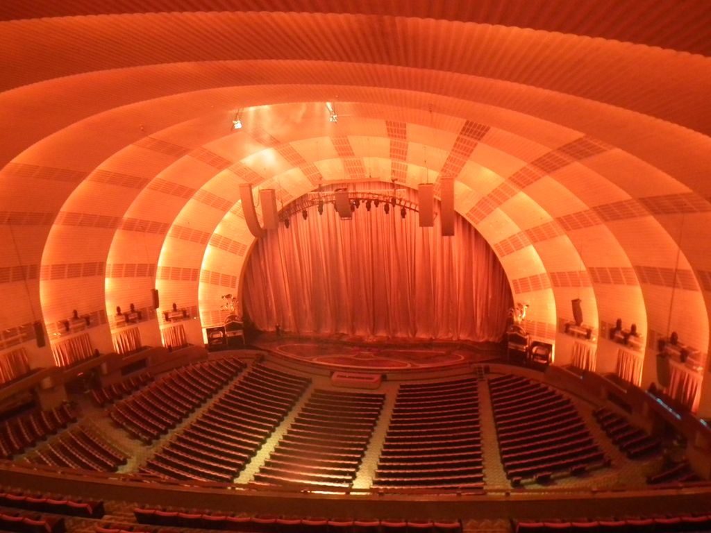 Illuminated concentric ceiling bands curve over rows of seats and aisles facing a curtained stage.