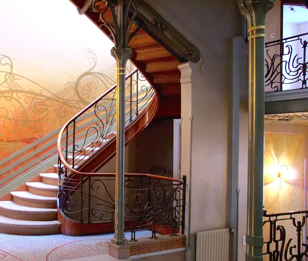 A curving staircase connects branching metal columns, flowing iron rails, wall patterns and mosaic flooring.