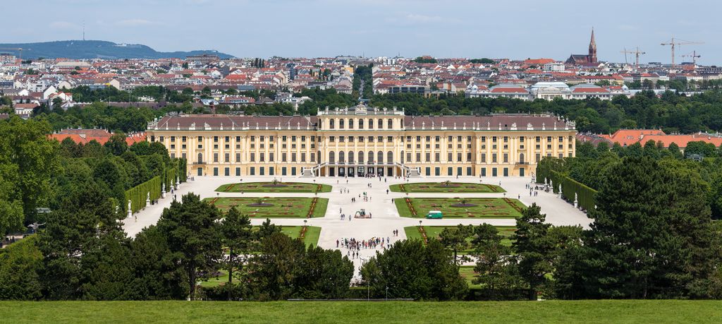Broad yellow palace aligned with a straight garden axis and paired formal flowerbeds, framed by trees with Vienna behind.