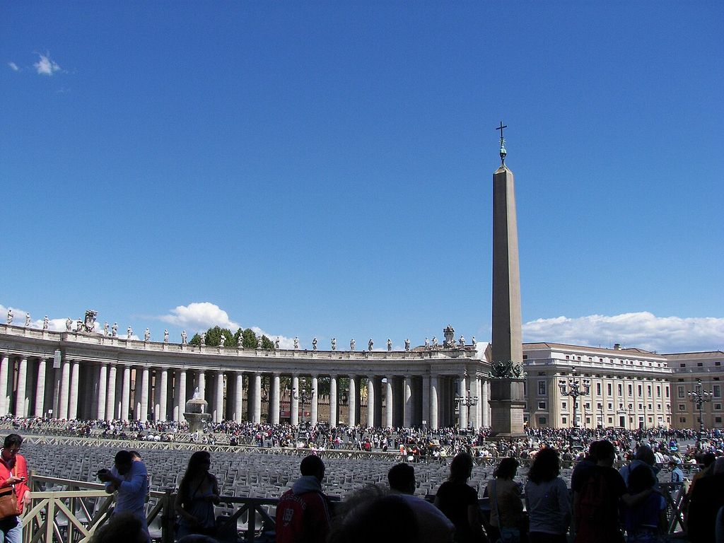 A curving colonnade frames an obelisk and crowds, with rows of temporary seating across Saint Peter’s Square.