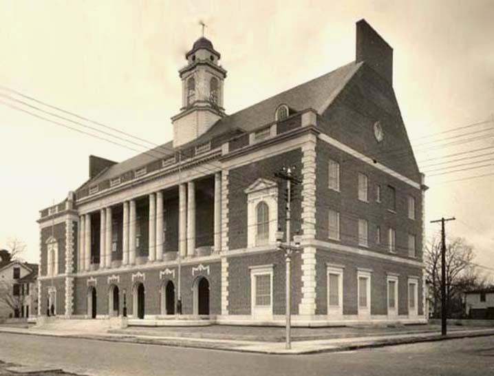 A large brick building with pale corner trim, paired columns and a cupola occupies a street corner.