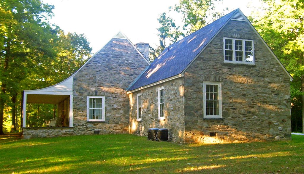 Low fieldstone wings with steep gabled roofs and white-framed windows meet beside an open porch overlooking a wooded lawn.