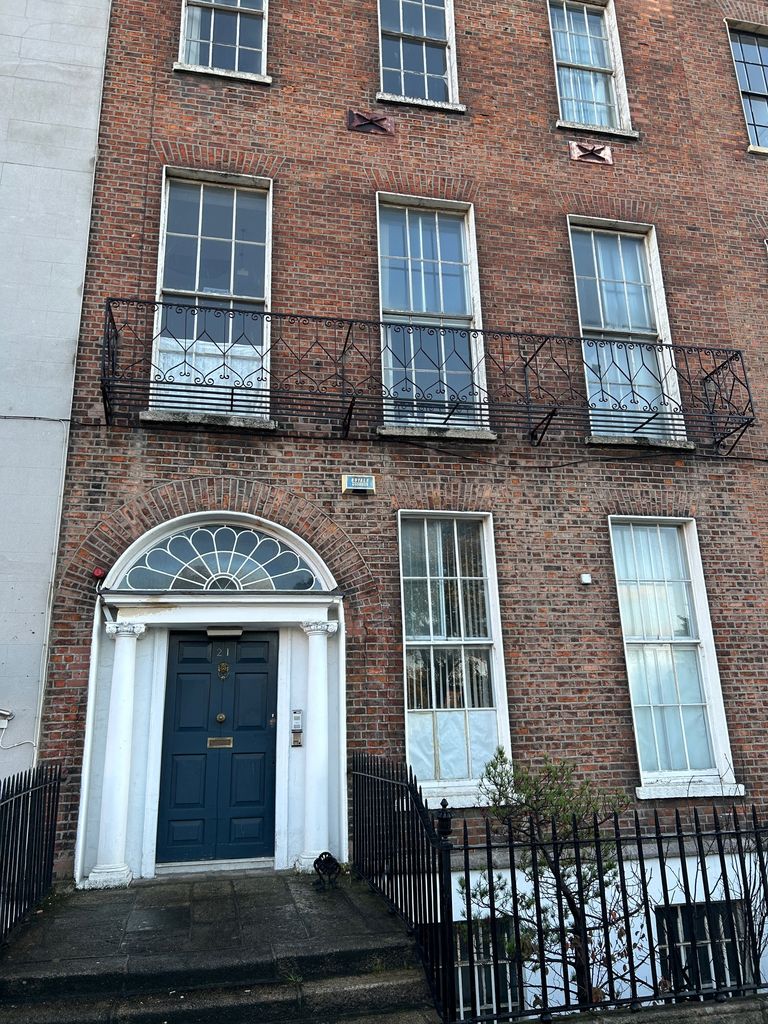 Red-brick façade with sash windows, an iron balcony, a blue door framed by Ionic columns and a fanlight, stone steps and basement railings.