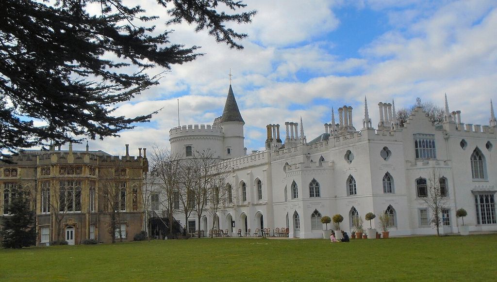 A pale house has battlements, pinnacles, pointed windows and a round tower beside a lawn.