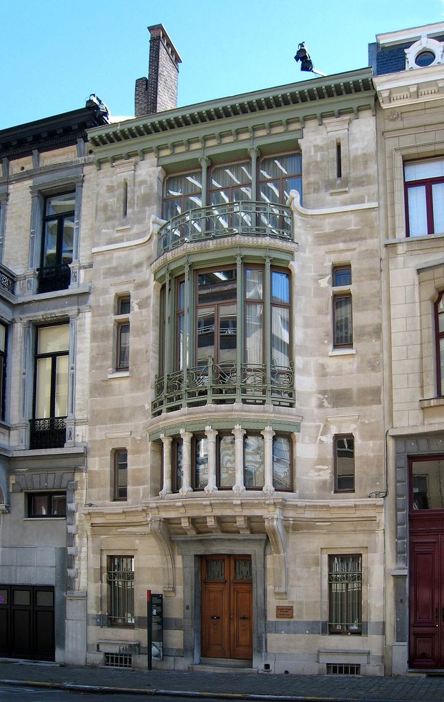 Curving iron railings and a projecting central window bay on the stone façade of Hôtel Tassel.