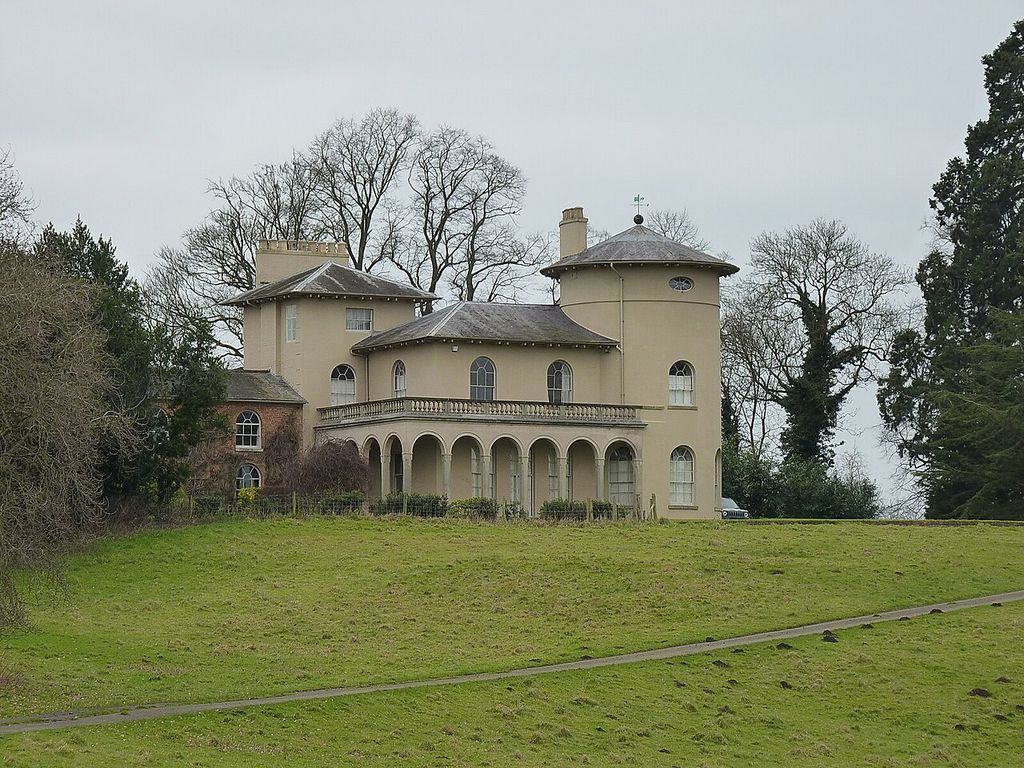 Round and square towers rise above a low-roofed villa with an arched loggia, set on a grassy slope.