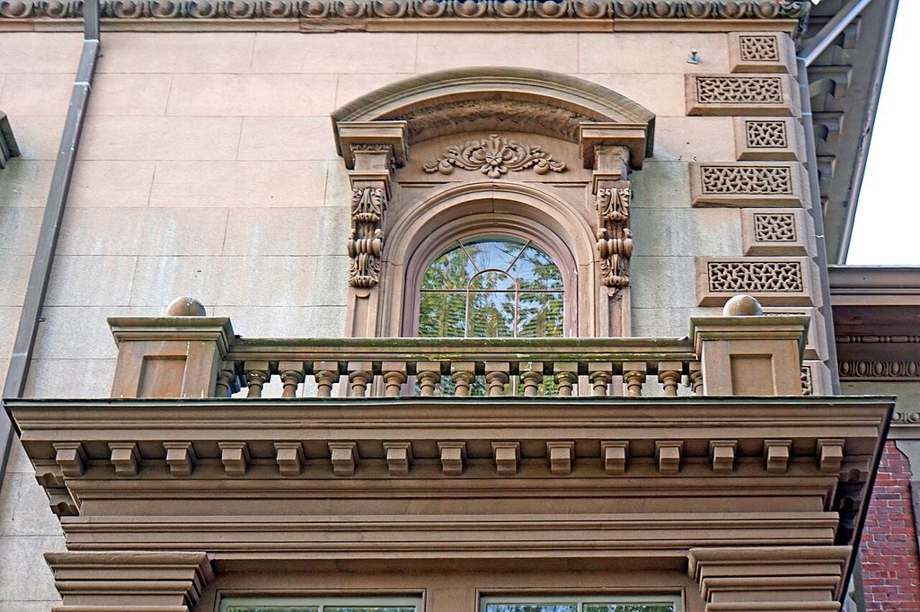 An arched carved window hood rises above a stone balustrade and bracketed balcony.
