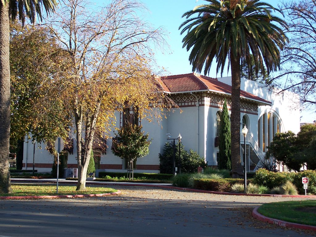 A hall with tall arched windows, a tiled roof and a geometric decorative band stands among mature trees.