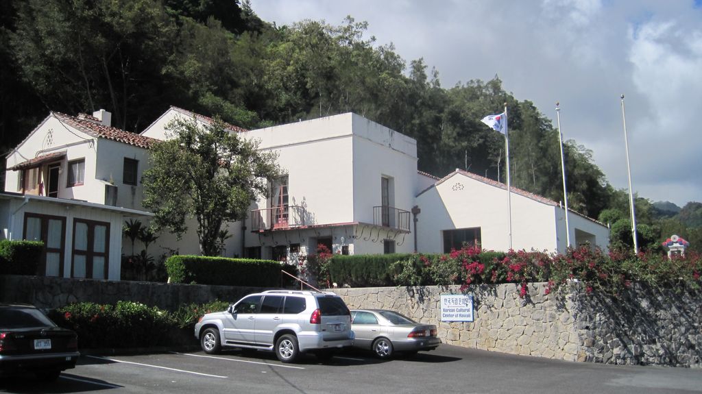 White house volumes with flat and tiled roofs step across a wooded slope above a stone retaining wall and parked cars.