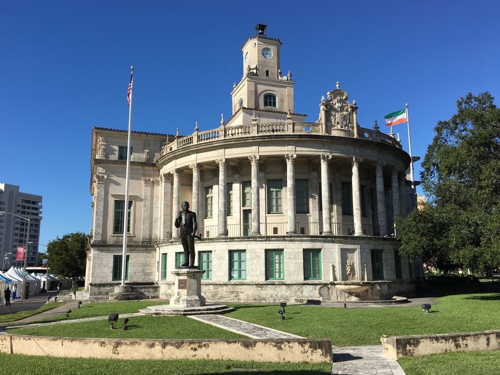 A curved colonnade and balustrade wrap around City Hall below its clock tower, with a lawn and fountain in front.