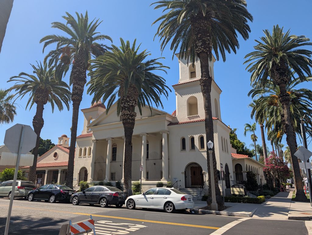 A church portico and paired towers rise behind tall palms, with tiled-looking red roofs, a side arcade and parked cars.