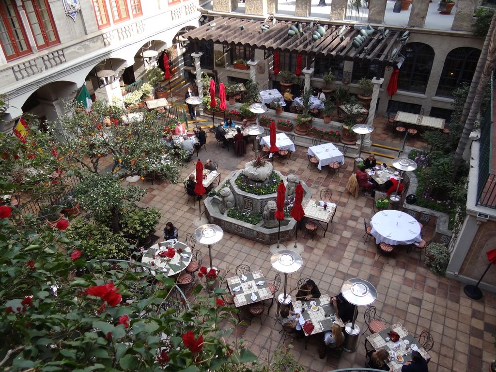Dining tables and a fountain occupy an enclosed hotel court below upper-level openings, arcades, planting and a pergola.