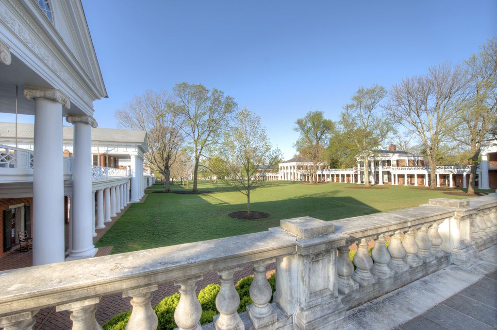 A stone balustrade overlooks a green lawn edged by low colonnades and larger classical pavilion fronts, with trees between them.