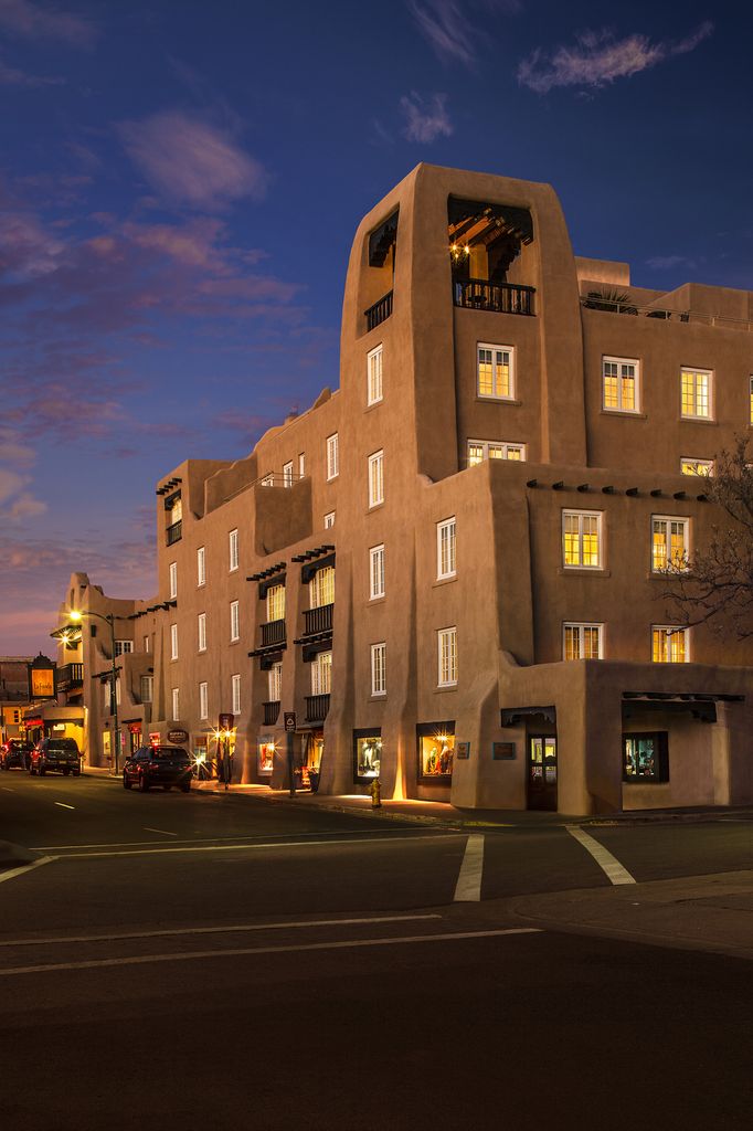 An illuminated hotel corner tower rises above stepped rooflines, small balconies and street-level shops at dusk.