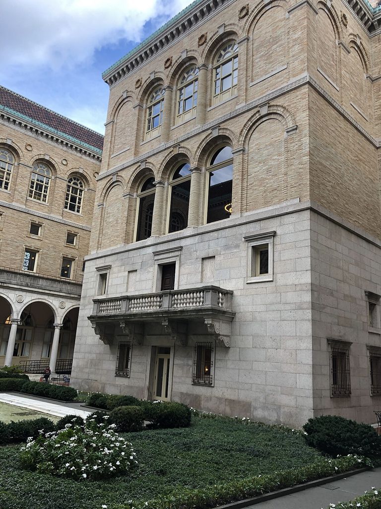 Stone lower walls, brick upper storeys and rows of arches enclose a planted courtyard corner.