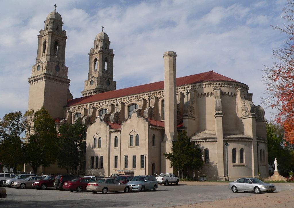 Twin domed towers rise behind a long red roof, shaped side-wall details and a curved apse above parked cars.