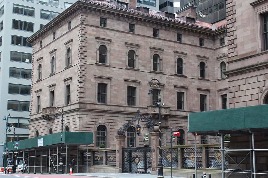 A pale masonry façade has a rusticated base, differently framed windows on successive floors and a deep cornice.