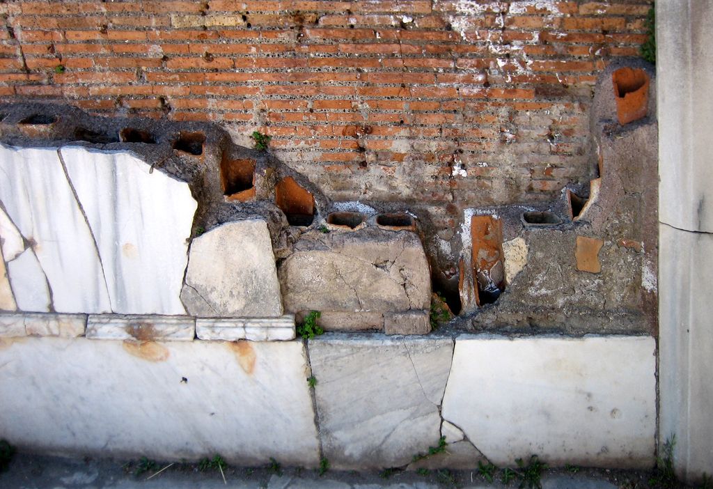 Open terracotta heating tubes exposed between a brick wall and fragmented pale wall facing at Ostia’s Terme del Foro.