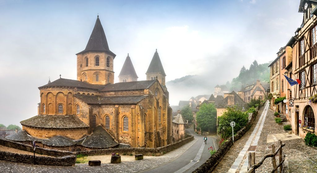 Layered stone apse and chapel roofs beneath church towers at Conques, beside a sloping village street in morning mist.
