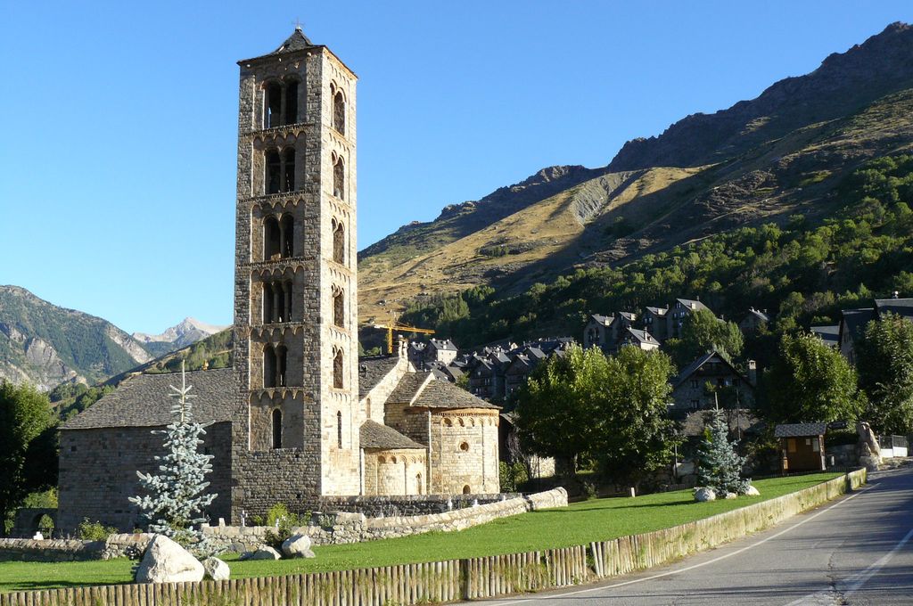 Tall stone bell tower with stacked arched openings beside the low roof and apses of Sant Climent, backed by mountain slopes and village buildings.