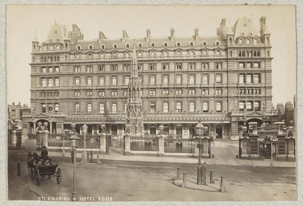 A long hotel façade has rows of dormers and tall roofed end pavilions, with the Queen Eleanor Memorial Cross and horse-drawn traffic in front.