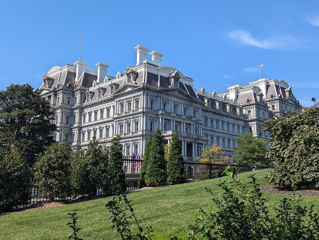 Repeated mansard pavilions, dormers and paired columns rise above gardens and a fence.