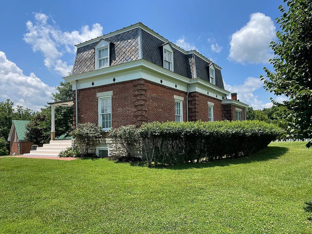A brick lodge has a steep slate mansard with dormer windows above a limestone base and clipped hedges.