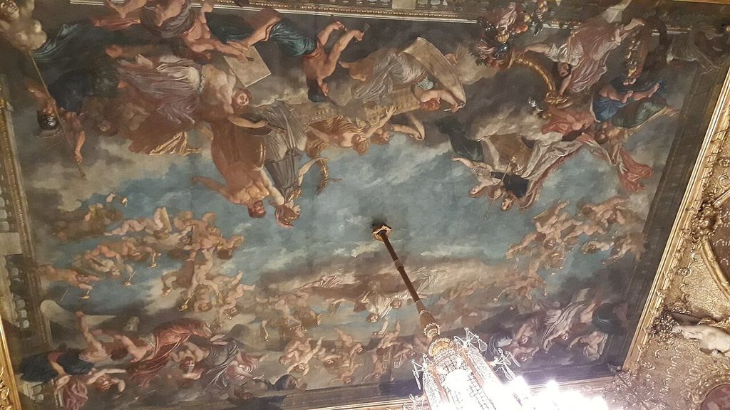 Numerous painted figures fill a large ceiling within an elaborate gilded frame, with a chandelier entering the lower edge.