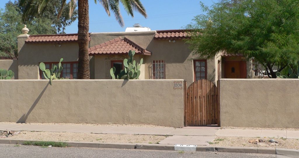 A low stucco house with uneven parapets and red tile-like roof strips stands behind a wall, wooden gate and palms.